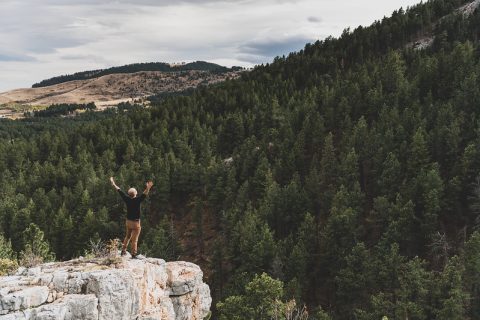 Exhilarated hiker celebrates atop a rugged cliff overlooking dense pine forests and rolling hills near Story, Wyoming — a serene escape for adventure seekers.