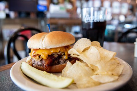 A juicy cheeseburger with bacon on a plate, served with potato chips and a pickle spear, with a soda in the background, ready to be enjoyed at a diner.