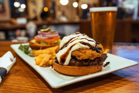 A close-up of a gourmet burger topped with melted cheese and sauce, served with fries on a plate, accompanied by a cold beer in a tall glass, set on a restaurant table.