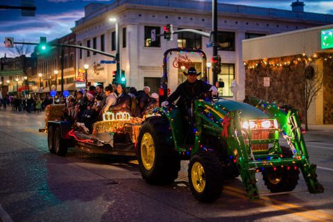 A decorated tractor adorned with colorful lights pulls a trailer full of people through a city street at dusk. The passengers are bundled up against the evening chill, enjoying a festive hayride. The city lights in the background add to the holiday atmosphere as the tractor's headlights shine brightly on the asphalt.