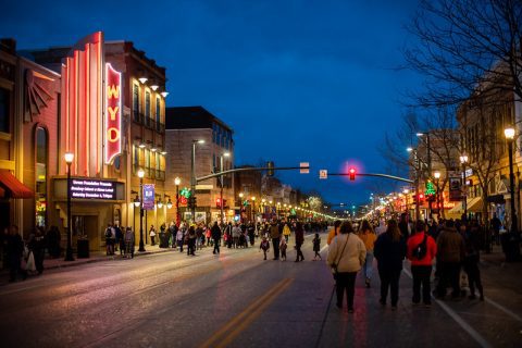An early evening shot of a bustling city street as twilight sets in. The neon marquee of the WYO Theater shines brightly, providing a warm welcome to the evening crowd. The holiday lights and the lively atmosphere of families and friends walking create a festive urban scene.
