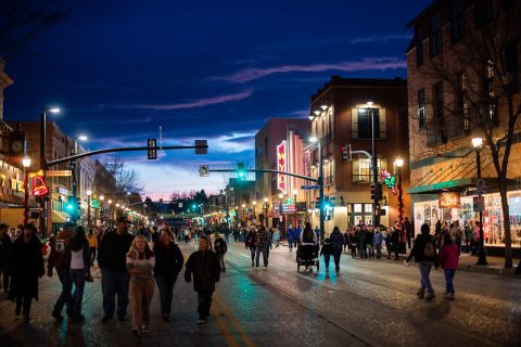 A vibrant evening scene on a busy downtown street, with pedestrians walking on the road lit by the soft glow of streetlights and storefronts. Holiday decorations add a festive touch, and the twilight sky above fades from blue to the deep indigo of approaching night.