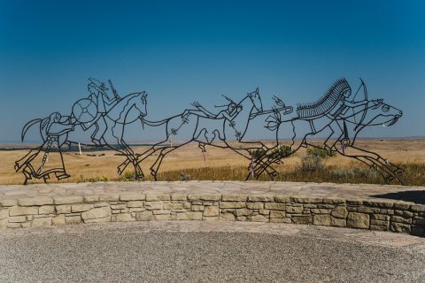 An outdoor metal sculpture depicting horsemen in motion, set against the expansive plains under a clear sky.