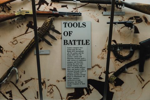 Historical firearms and weaponry displayed on a backdrop with a descriptive panel titled 'TOOLS OF BATTLE', detailing the arms used by Custer's troops.