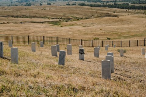 White headstones dot a dry grassy field in a solemn cemetery, with rolling hills stretching into the distance.
