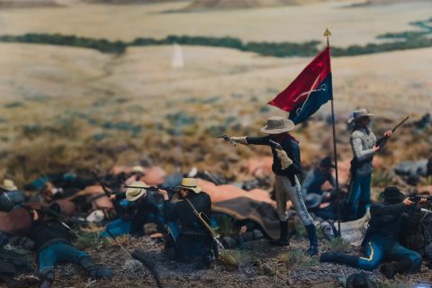 Miniature diorama depicting a historical battle scene with soldiers in combat, a flag bearer holding a regimental standard, against a painted prairie backdrop.