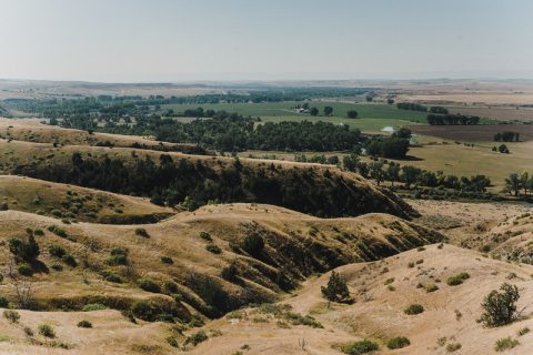 A sweeping landscape of rolling hills with sparse vegetation transitions into lush farmland in the distance.