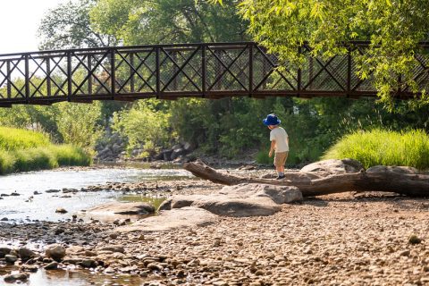 A child explores a pebbled creek under a rustic bridge at Kendrick Park, a cherished activity and top thing to do for families in Sheridan, Wyoming.