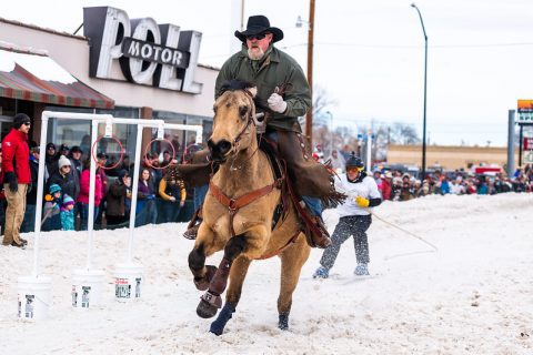 A cowboy on horseback speeds through the snow in Sheridan, WY, illustrating the excitement of skijoring events.