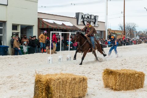 A horse and rider skillfully navigate a skijoring course in Sheridan, WY, leaping over a jump as spectators watch the exciting competition.