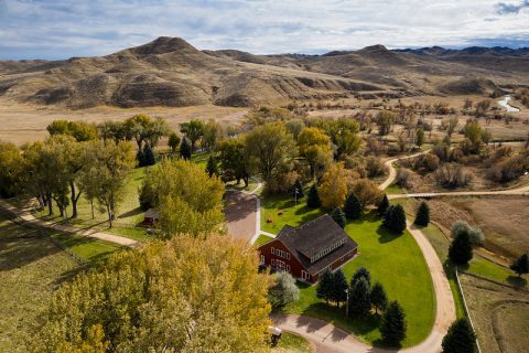 Aerial view of Ucross, Wyoming, featuring a striking red barn amid lush greenery, with winding rivers and rolling hills in the background, capturing the essence of rural beauty.